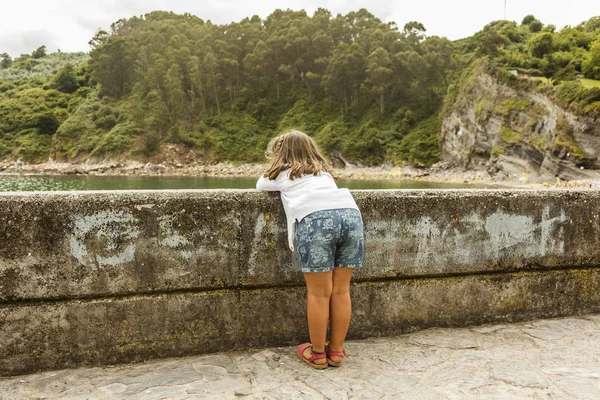 Vista trasera chica viendo el paisaje marino descansando en la pared. Asturias . — Foto de Stock