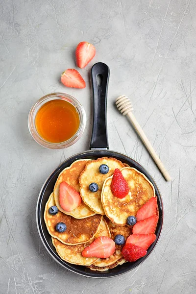 Pancakes in a frying pan with strawberries and blueberries and honey, gray background, copy space