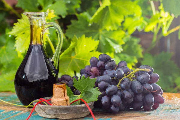 黑色的老香醋在一个玻璃水罐与新鲜的红色葡萄在绿色葡萄园背景 — 图库照片