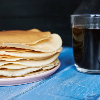 Mavi ahşap bir masada, bir tabak sade kahvenin üzerinde bir yığın krep.