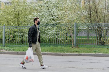 Koruyucu maskeli bir adam elinde poşetle sokakta yürüyor.