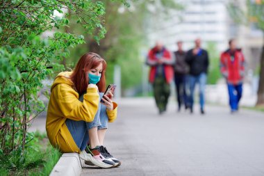 Kız kaldırımda oturuyor koruyucu bir maskeyle ve elinde bir telefonla.