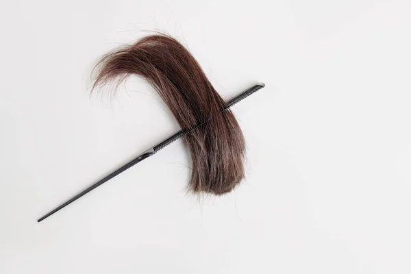 她棕色的 光滑的头发的剪下来的尖端被交叉在一个浅灰背景的梳子上 白色背景上的一缕剪下来的深色头发和一把梳子 — 图库照片