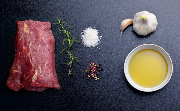 Rauwe biefstuk, zout, peper, knoflook, rozemarijn, olijfolie op het zwarte bord, achtergrond. — Stockfoto