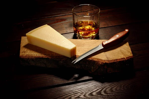 Queso parmesano en una tabla de madera con cuchillo y vaso de whisky en el segundo plan con hielo —  Fotos de Stock