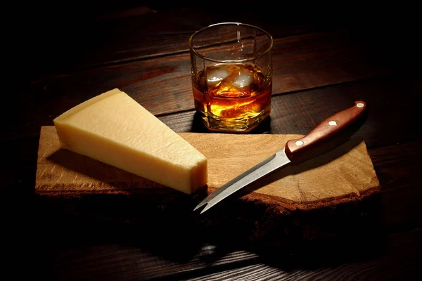 Queso parmesano en una tabla de madera con cuchillo y vaso de whisky en el segundo plan con hielo —  Fotos de Stock