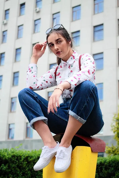 Retrato de moda de una joven chica hipster bonita con pelos marrones oscuros que usan tirantes elegantes para pantalones, gafas y camisa con una divertida ubicación de cereza estampada en un vertedero amarillo. — Foto de Stock