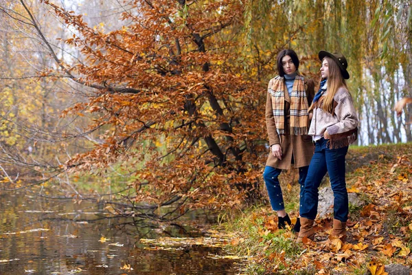 Moda piękny portret dwóch pięknych kobiet, stojąc na brzegu jesień jezioro krajobraz. Modny płaszcz indian i czapkę z szalikiem. Moda Jesień koncepcja. Zbliżenie — Zdjęcie stockowe