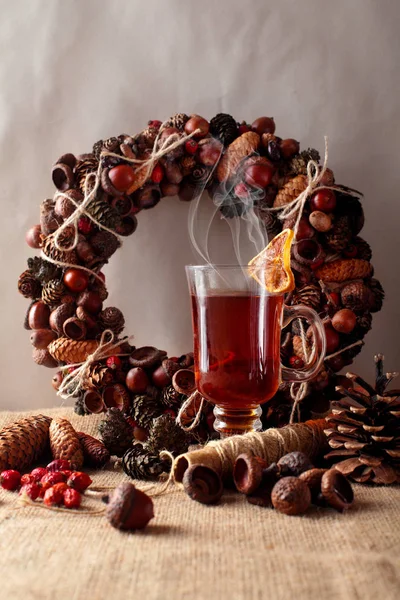 A cup or glass of the hot spicy tea drink with dried lemon.Christmas wreath of arcorns and cones on second plan, threads and dog rose.Warm athmosphere — стоковое фото