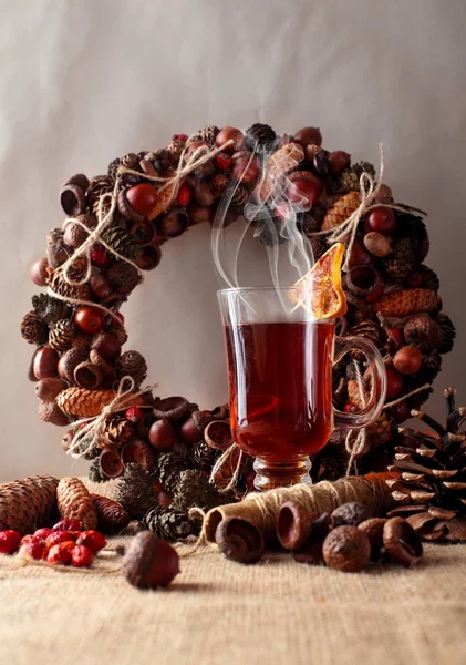 A cup or glass of the hot spicy tea drink with dried lemon.Christmas wreath of arcorns and cones on second plan, threads and dog rose.Warm athmosphere — стоковое фото