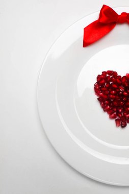 Nar taneleri bir kalp şeklinde beyaz levha ve Şerit'in kırmızı yay. Beyaz arka plan üzerinde. Sevgililer günü için arka plan. Minimalism.Copy alanı. Portre