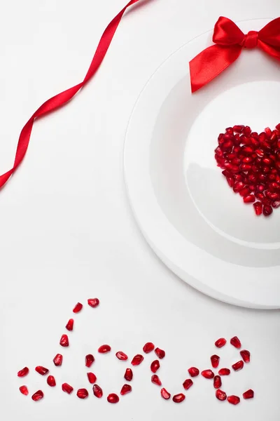 Pomegranate grains in heart shape on white plate and red bow of ribbon.Word love of Pomegranate grains .On white background.Background for valentine's day.Minimalism.Copy space.Closeup — Stock Photo, Image