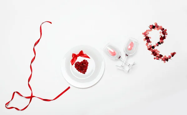 Love word of the red ribbon, white plate with Pomegranate seed in shape heart, glasses of wine, petals of dried roses on white background on red napkin line background.Valentine 's day creative backgr — стоковое фото