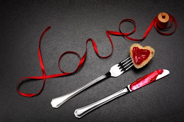 Iscrizione, parola amore per il nastro di raso rosso e cuore di pane tostato con marmellata rossa su forchetta e coltello con marmellata.San Valentino sullo sfondo del giorno.Love concept.Sullo sfondo pietra scura.Creativo.Love backgound — Foto Stock