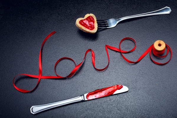 Inscription, word love of red satin ribbon and heart of toast bread with red jam on fork and knife with jam.Valentine day background.Love concept.On dark stone background.Creative.Love background