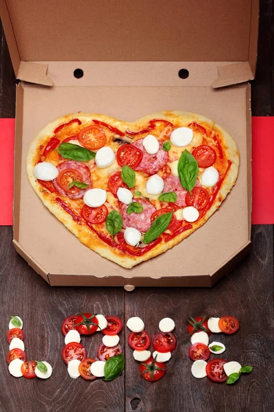 Pizza em forma de coração com salame, tomates, manjericão e mussarela. Em caixa de papelão contra um fundo marrom de madeira escura. Inscrição amor de tomates mussarela e manjericão Vista superior. Valenti criativo — Fotografia de Stock