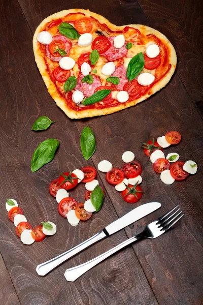 Pizza in shape heart with salami, tomatoes, basil and mozzarella with fork and knife.Against a dark wooden brown background. Inscription love of mozzarella tomatoes and basil .Top View. Creative valen