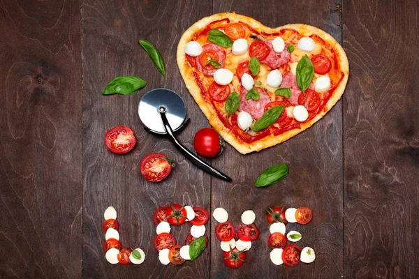 Pizza in shape heart with salami, tomatoes, basil and mozzarella with knife for pizza.Against a dark wooden brown background. Inscription love of mozzarella tomatoes and basil .Top View. Creative vale