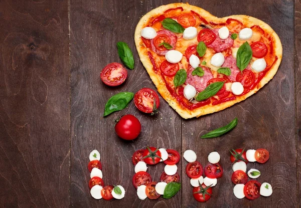 Pizza in shape heart with salami, tomatoes, basil and mozzarella with knife for pizza.Against a dark wooden brown background. Inscription love of mozzarella tomatoes and basil .Top View. Creative vale