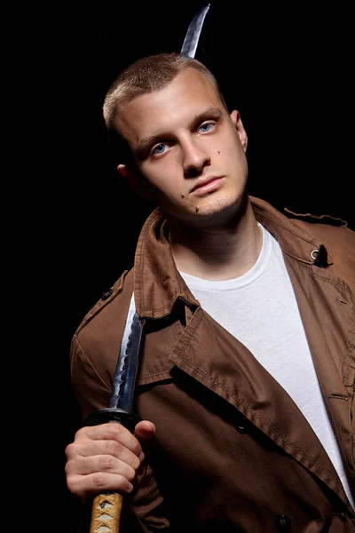 Young man dressed in a brown coat and white t-shiert, holding a katana.Modern samuria.Fashion portrait.Urban warior.Dramatic light — Stock Photo, Image