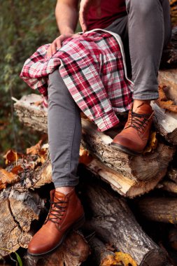 Kahverengi deri çizmeli erkek ayağı, gri kot pantolon ve bacağında kırmızı gömlek yakacak odun yığınına sokuluyor.
