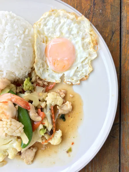Mix of fried vegetables, Cauliflower, Carrot, Sugar Pea with seafood with rice and fried egg in white plate on wooden background. Thai style food.