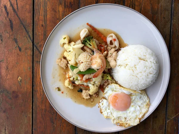 Mix of fried vegetables, Cauliflower, Carrot, Sugar Pea with seafood with rice and fried egg in white plate on wooden background. Thai style food.