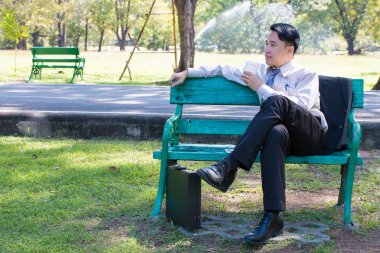 Yeşil Ahşap bankta oturan ve bir beyaz kağıt bardak su, kahve veya çay için elinde tutan genç iş adamı almak uzakta ve içme, yeşil orman park açık havada çalıştıktan sonra rahatlatıcı.