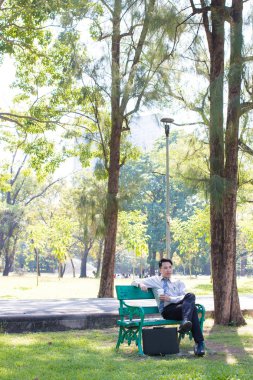 Yeşil Ahşap bankta oturan ve bir beyaz kağıt bardak su, kahve veya çay için elinde tutan genç iş adamı almak uzakta ve içme, yeşil orman park açık havada çalıştıktan sonra rahatlatıcı.