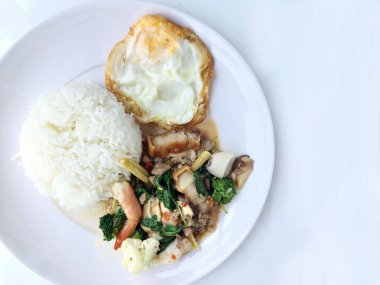 Kızarmış gevrek domuz eti ilave edin, karides, Tay kutsal fesleğen, chilies, sarımsak ve soya sosu ile deniz ürünleri karıştırın. kızarmış yumurta stil tepesi beyaz arka plan üzerinde beyaz tabak pirinç ve Tay dili ile.