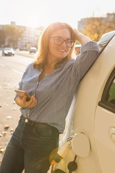 Mujer joven está de pie cerca del coche eléctrico y la celebración de teléfono inteligente —  Fotos de Stock