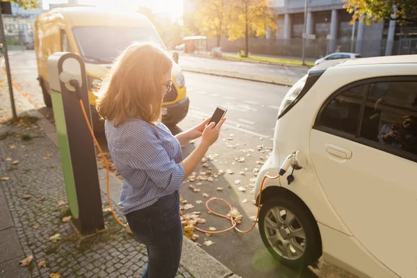 Jovem mulher está de pé perto do carro elétrico e segurando smartphone — Fotografia de Stock