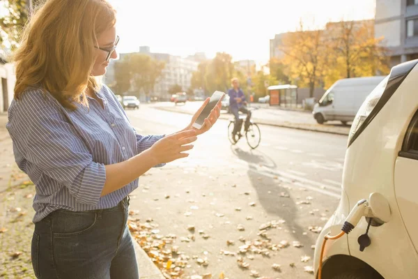 Młoda kobieta jest stojący w pobliżu samochód elektryczny i trzymając smartfon — Zdjęcie stockowe