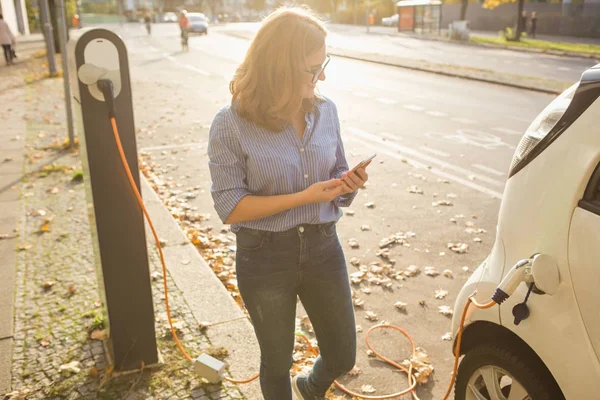 Mujer joven está de pie cerca del coche eléctrico y la celebración de teléfono inteligente — Foto de Stock
