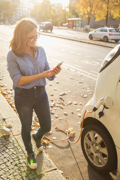 Jovem mulher está de pé perto do carro elétrico e segurando smartphone — Fotografia de Stock
