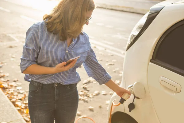 Jovem mulher está de pé perto do carro elétrico e segurando smartphone — Fotografia de Stock