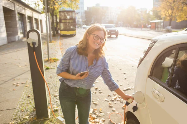 Jovem mulher está de pé perto do carro elétrico e segurando smartphone — Fotografia de Stock