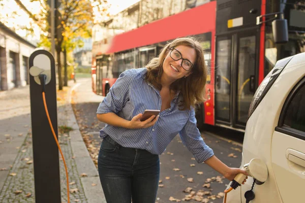 Junge Frau steht in der Nähe des Elektroautos und hält Smartphone — Stockfoto