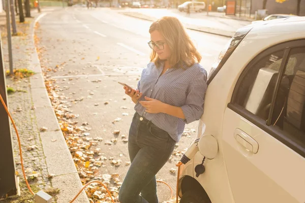 Young woman is standing near the electric car and holding smartphone — Stock Photo, Image