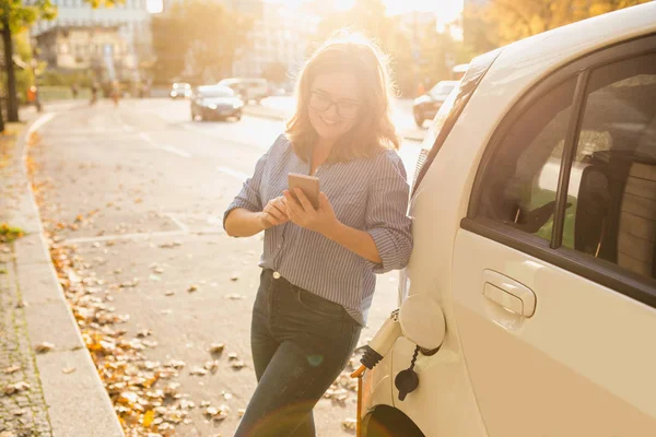 Młoda kobieta jest stojący w pobliżu samochód elektryczny i trzymając smartfon — Zdjęcie stockowe