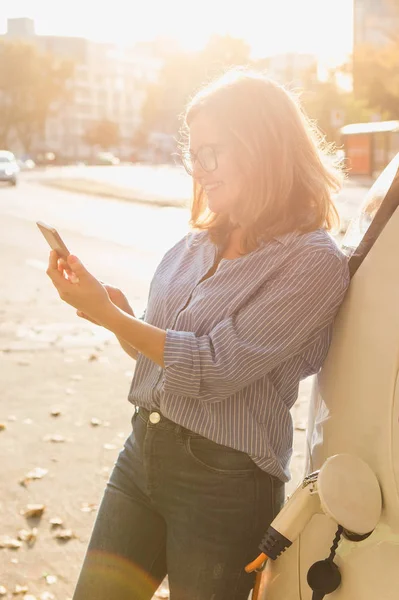 Fiatal nő közelében az elektromos autó, és a gazdaság smartphone — Stock Fotó