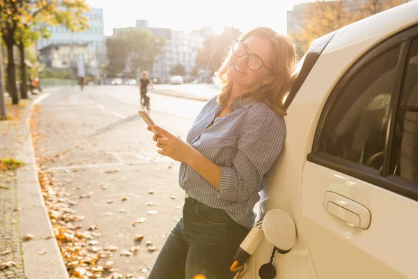 Młoda kobieta jest stojący w pobliżu samochód elektryczny i trzymając smartfon — Zdjęcie stockowe