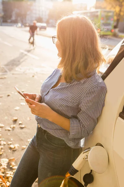 Jonge vrouw is permanent in de buurt van de elektrische auto en bedrijf smartphone — Stockfoto