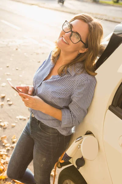 Jonge vrouw is permanent in de buurt van de elektrische auto en bedrijf smartphone — Stockfoto