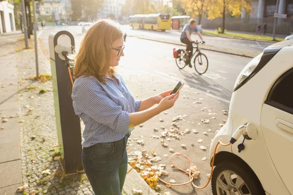 Mujer joven está de pie cerca del coche eléctrico y la celebración de teléfono inteligente — Foto de Stock