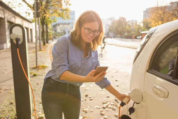 Jovem mulher está de pé perto do carro elétrico e segurando smartphone — Fotografia de Stock