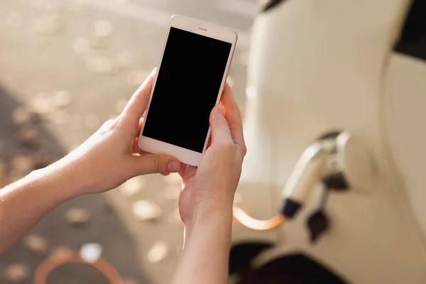 Young woman is standing near the electric car and holding smartphone — Stock Photo, Image