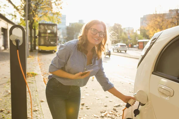 Mujer joven está de pie cerca del coche eléctrico y la celebración de teléfono inteligente —  Fotos de Stock