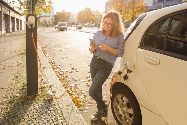 Молодая женщина стоит возле электромобиля и держит в руках смартфон — стоковое фото