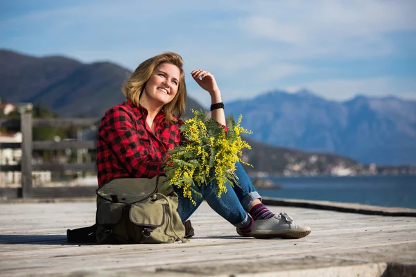 Happy girl voyageur assis sur une jetée en bois et tient un bouquet de fleurs de mimosa dans les mains . — Photo